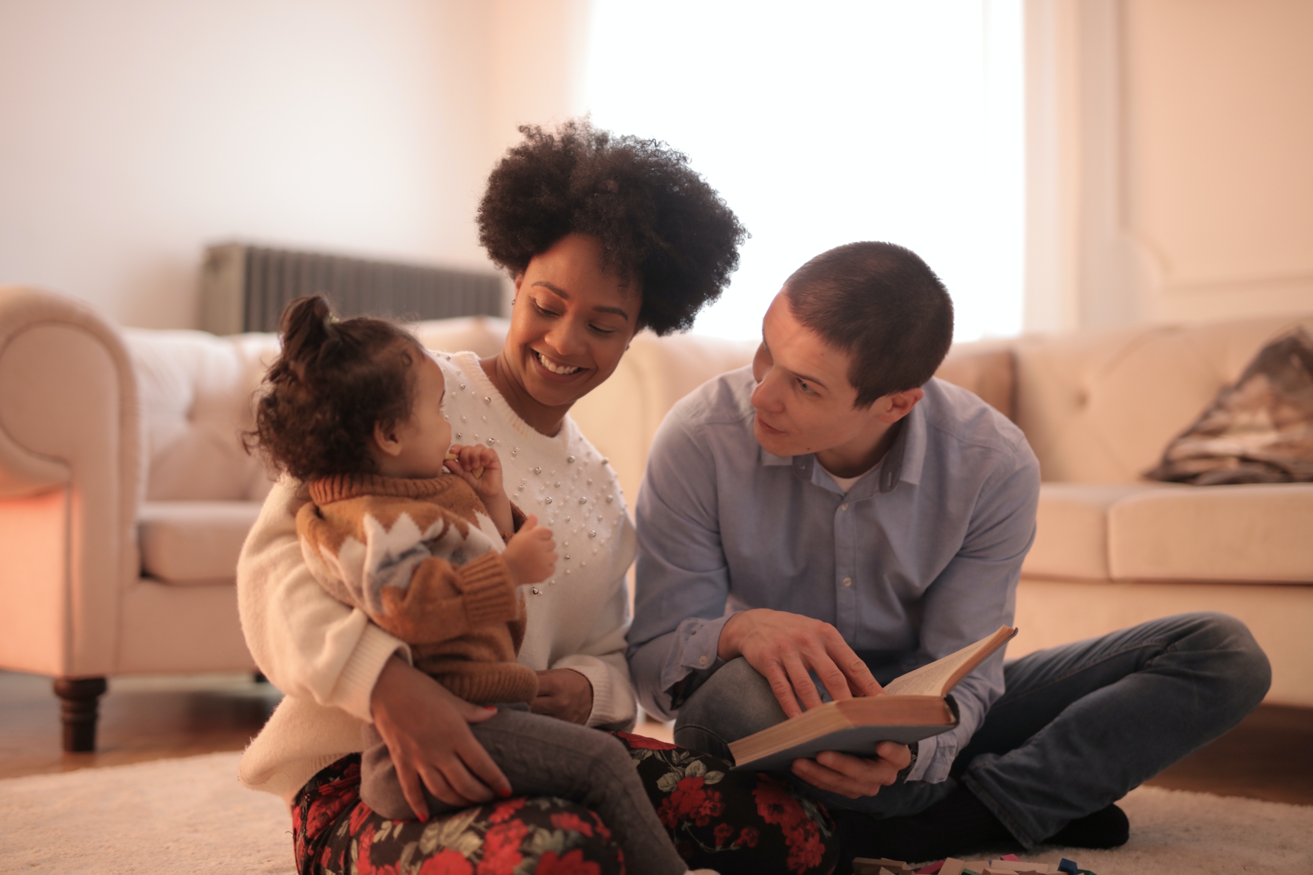 A woman, man, and toddler sitting on the floor reading a book, a good practice according to Child Care Aware of America